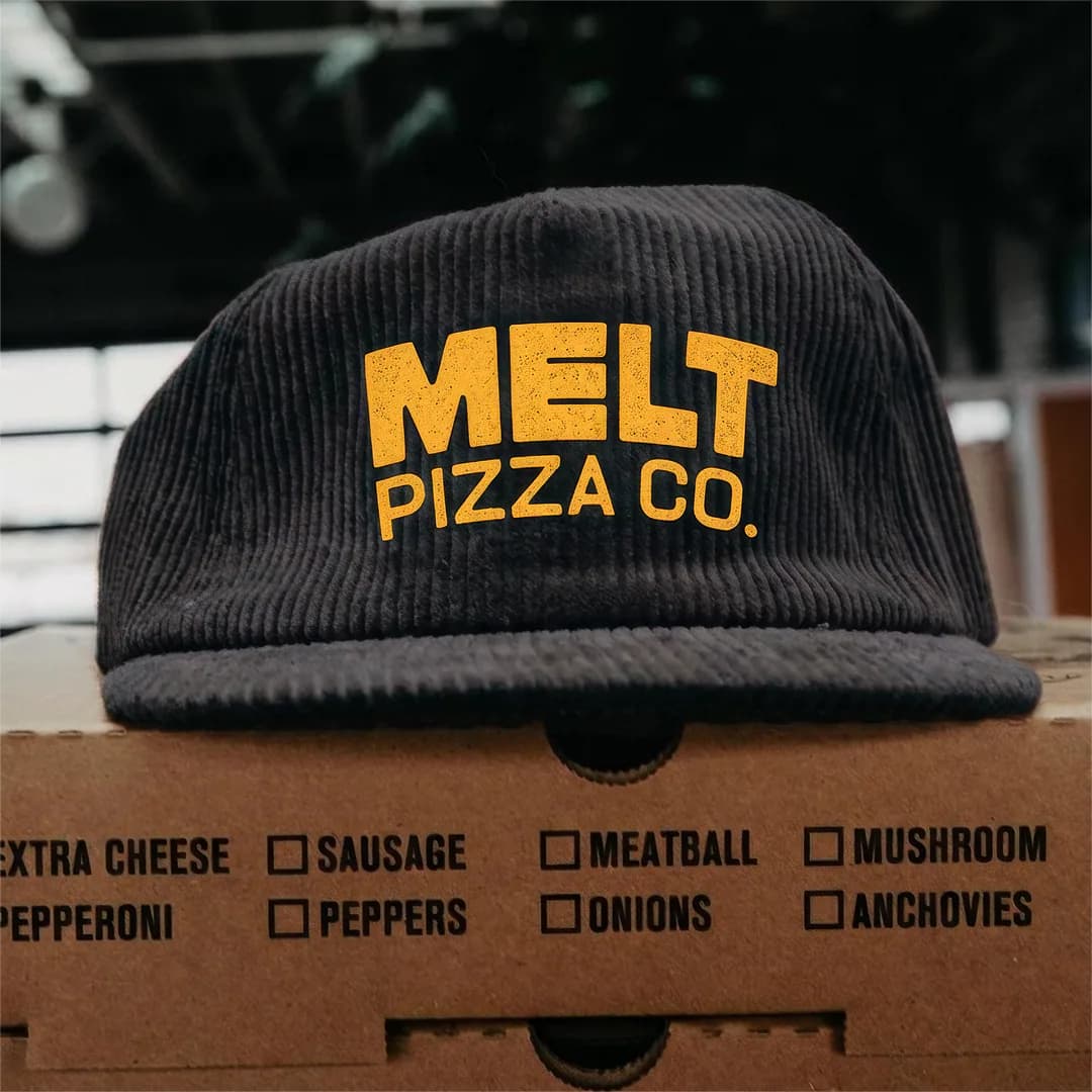 Melt Pizza Co branded corduroy cap on a pizza box topping checklist